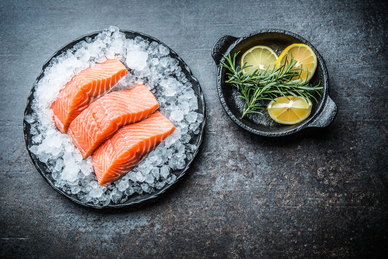 A delectable collection of three salmon filets on ice sitting next to a small bowl of lemon halves.